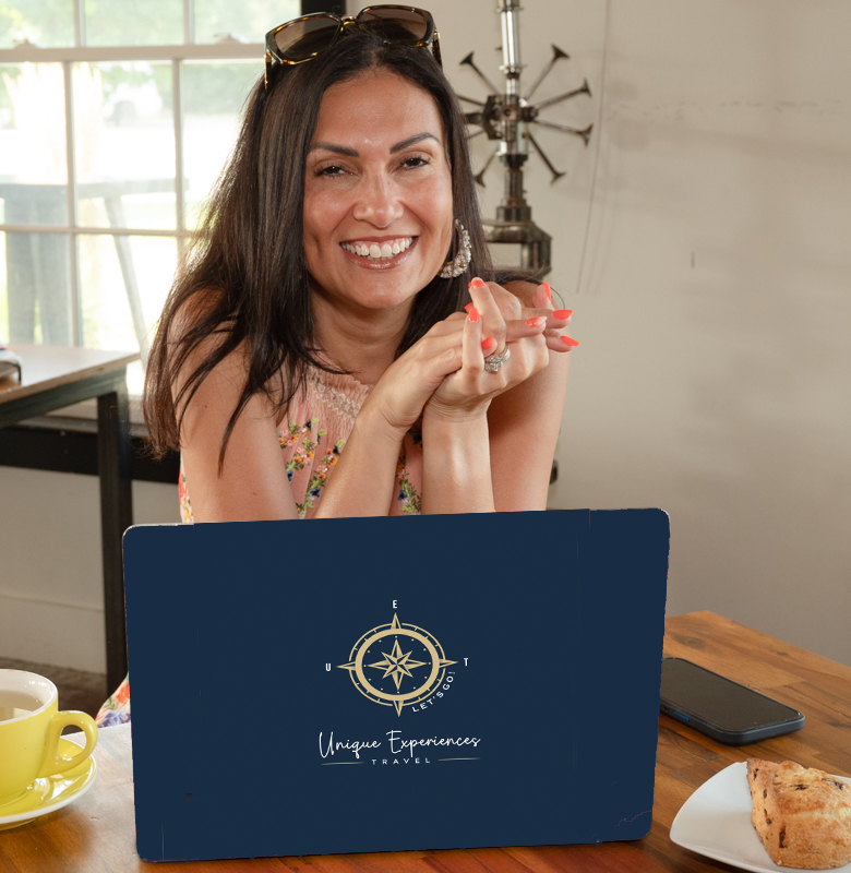 about us image - Unique Experiences Travel Smiling woman seated at a wooden table with a blue "Unique Experiences" laptop, coffee, and pastry, in a sunlit room with a decorative windmill and large window.