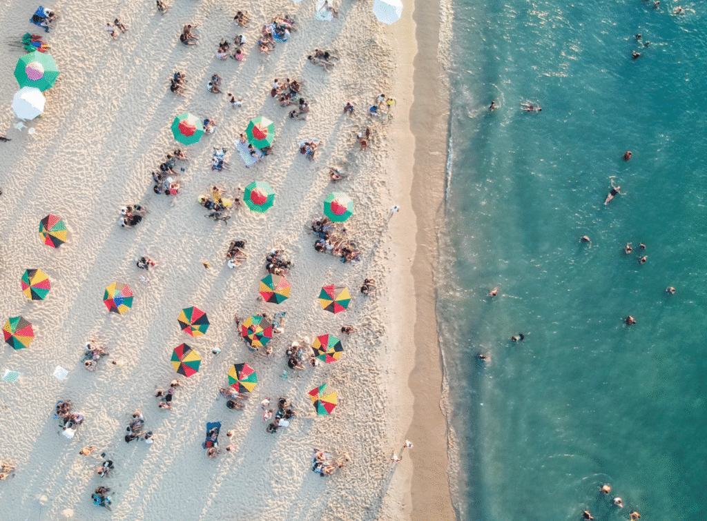 Aerial view of a crowded sandy beach with colorful umbrellas and groups of sunbathers near the shoreline and turquoise water to the right.