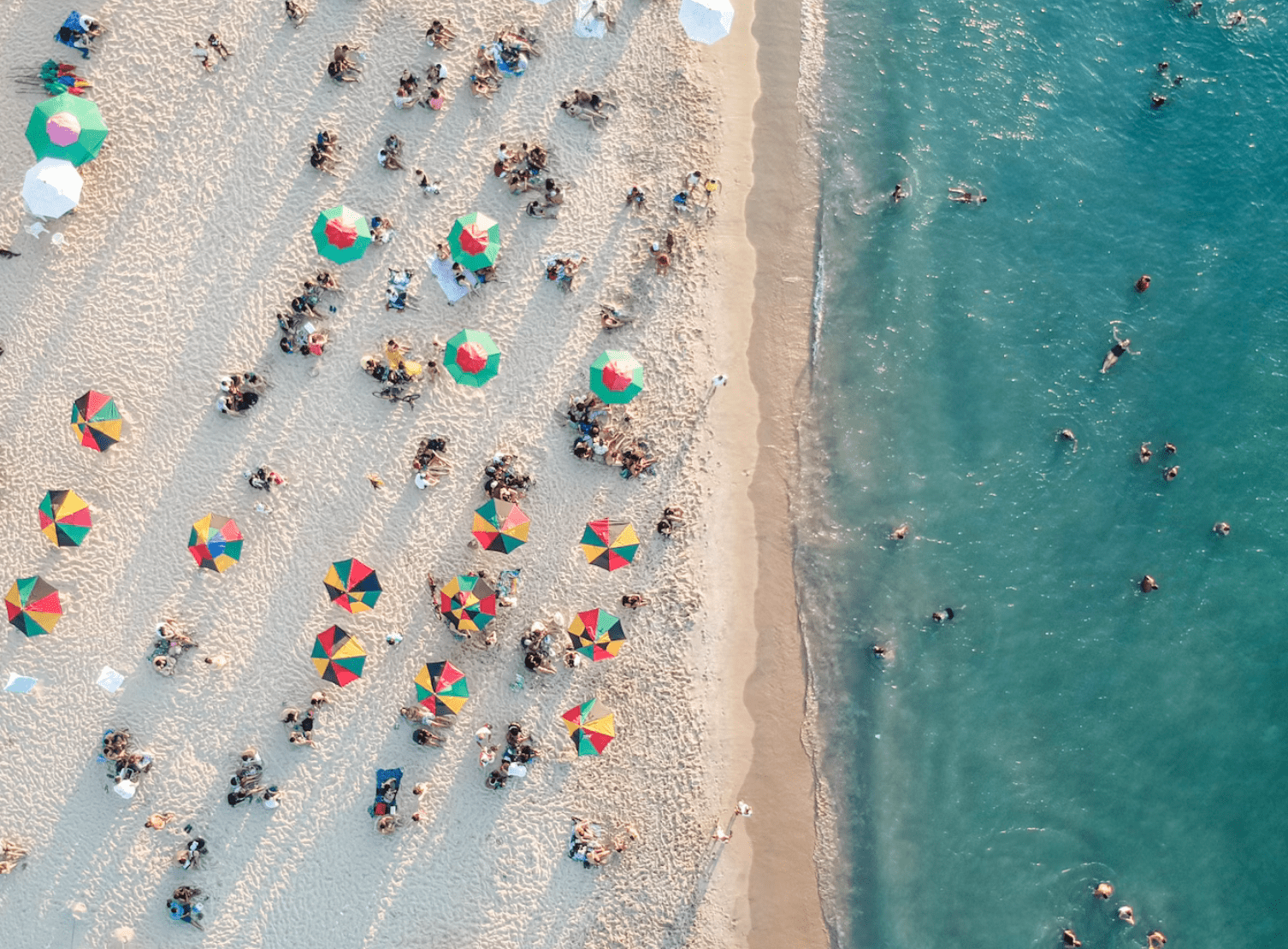Aerial view of a crowded sandy beach with colorful umbrellas and groups of sunbathers near the shoreline and turquoise water to the right.