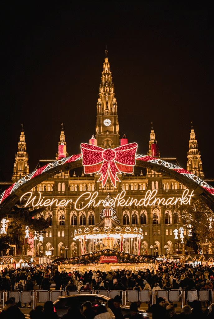 Night view of Vienna's Wiener Christkindlmarkt with a bright red bow arch and illuminated historic buildings, crowds bustling below.