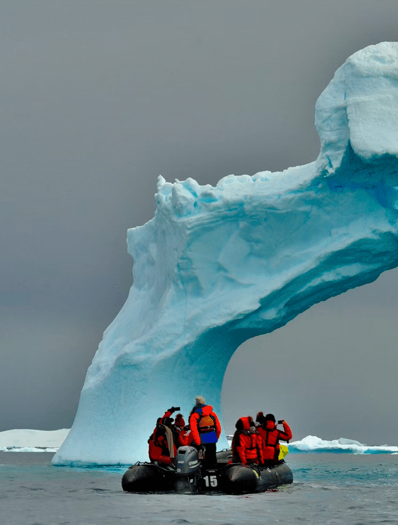 Group of explorers in a black inflatable boat wearing red life jackets under a towering blue iceberg arch in icy water.