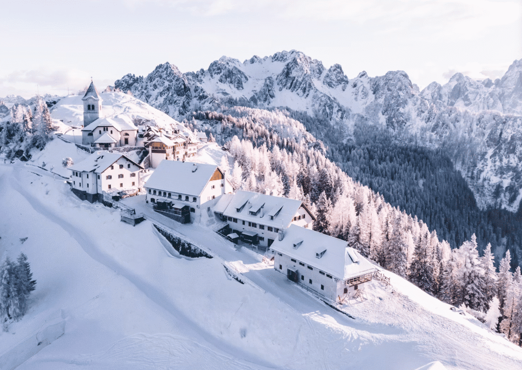 Snow-covered alpine village on a hillside with a church and wooden chalets amid snow-laden pine trees and towering mountains in the background.