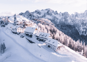 Snow-covered alpine village on a hillside with a church and wooden chalets amid snow-laden pine trees and towering mountains in the background.