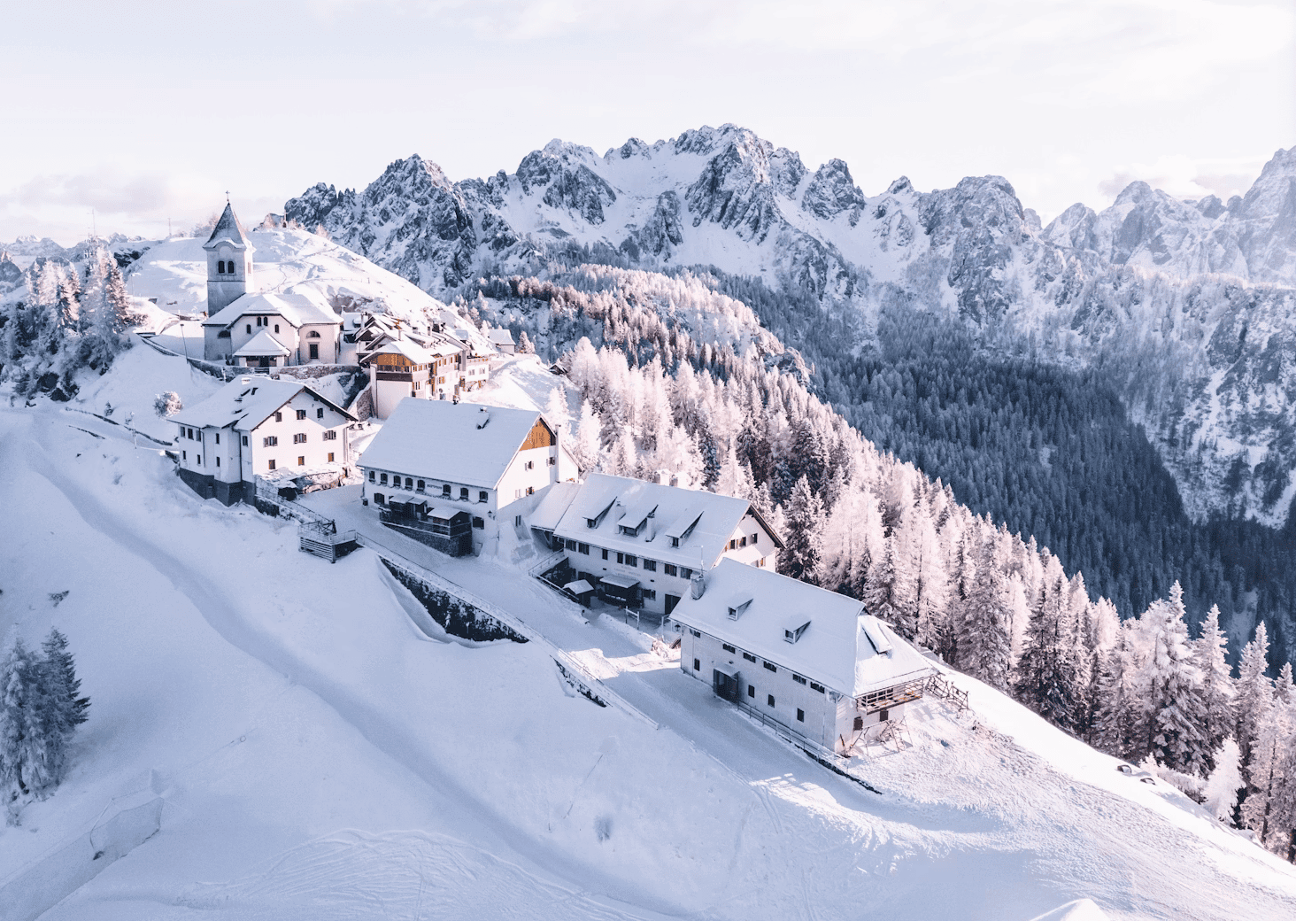 Snow-covered alpine village on a hillside with a church and wooden chalets amid snow-laden pine trees and towering mountains in the background.