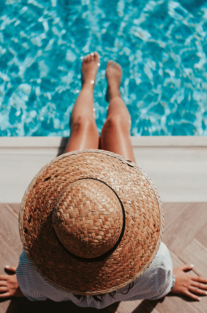 Person sitting at a pool edge with legs extended, wearing a woven straw hat, toes near the blue water.