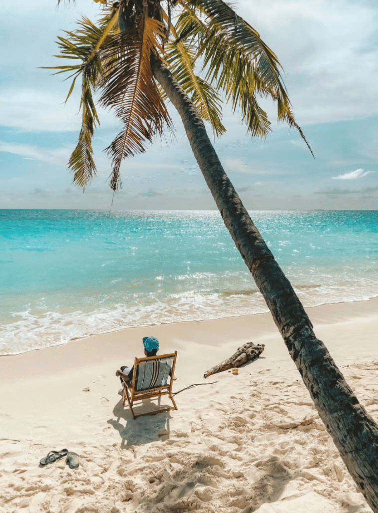Person relaxing in a wooden beach chair under a leaning palm tree on a white-sand tropical beach, turquoise sea in the background.