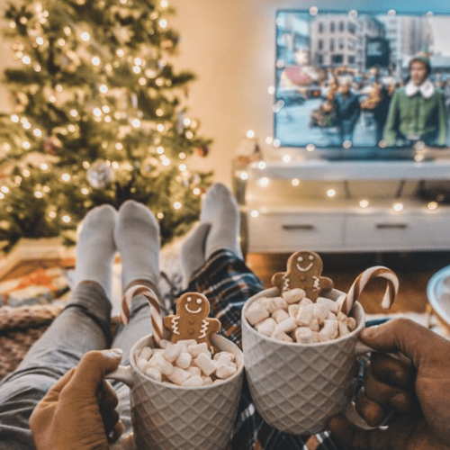 Couple relaxing in plaid pajamas with hot chocolate and gingerbread cookies, watching “Elf” by a Christmas tree—capturing cozy holiday moments and winter travel downtime.